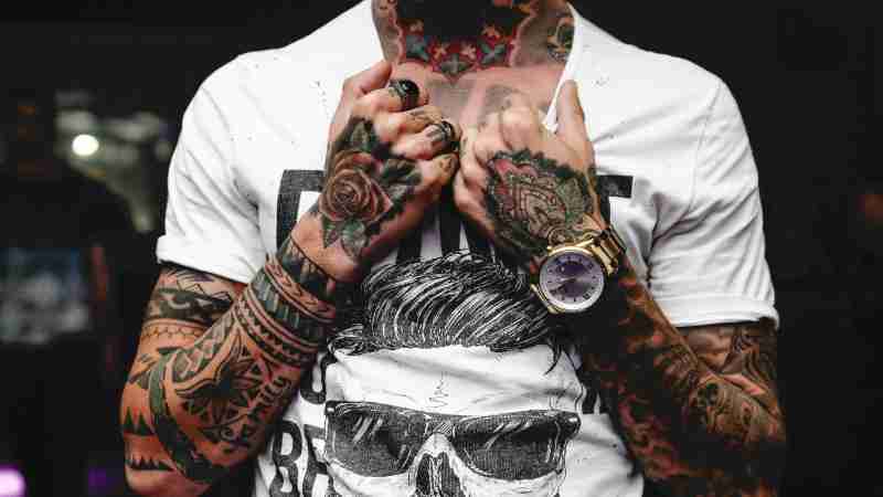 Close-up of tattooed arms and hands clutching a graphic t-shirt featuring skull art., tags: risico op - pexel